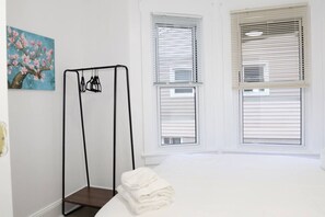 Une pièce comprenant un lit, un étendoir fixé au mur et deux fenêtres équipées de stores.