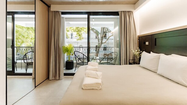 A modern hotel room with a large bed, a balcony with chairs, and a view of greenery. - Portal Arévalo in Palermo by Magno | 12 (Buenos Aires)
