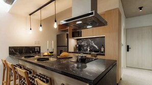 A modern kitchen with a central island, pendant lights, a stainless steel range hood, and wooden chairs. - Portal Arévalo in Palermo by Magno | 12 (Buenos Aires)