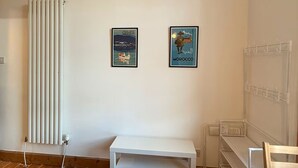 Ein Zimmer mit einem weißen Heizkörper, einer Sitzbank, einem kleinen Tisch und zwei gerahmten Postern an der Wand.
