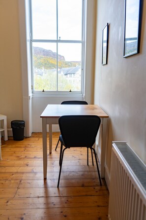 Ein Zimmer mit Holzboden, einem weißen Tisch, einem schwarzen Stuhl, einem Fenster mit Blick auf ein Gebäude und Berge sowie einer Heizung.