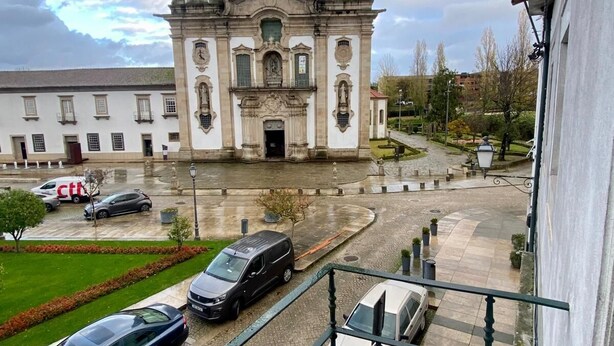 Vista de un edificio con dos torres destacadas y una entrada central, rodeado de una zona pavimentada con coches y un pequeño jardín.