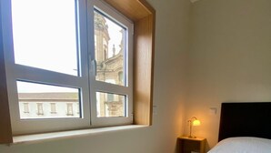 Habitación de hotel con una cama grande, ventana con vista a edificios, lámpara de noche y cabecero de madera.