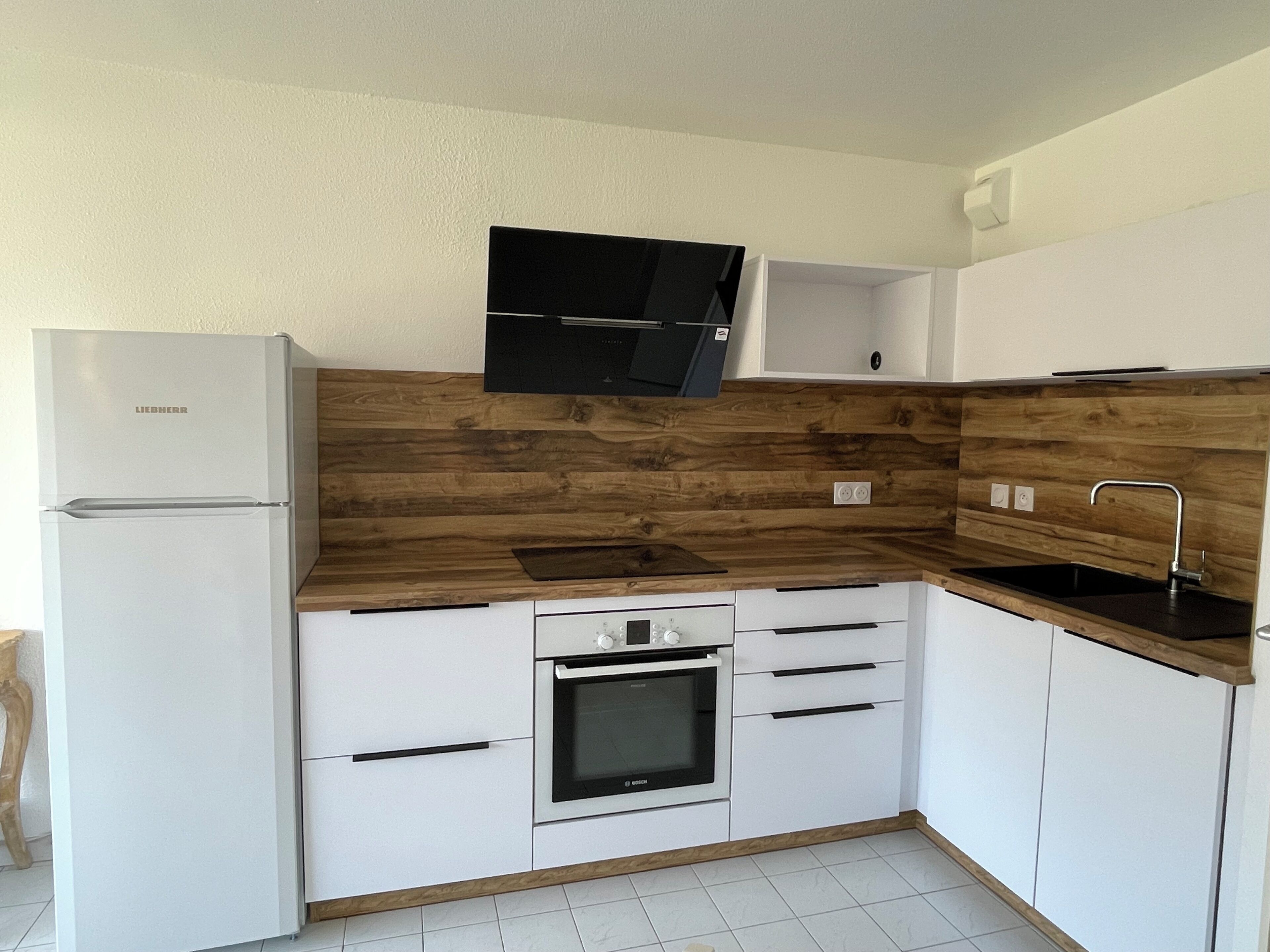 A kitchen with a built-in oven, a refrigerator, wooden backsplash, white cabinets, and a black countertop.