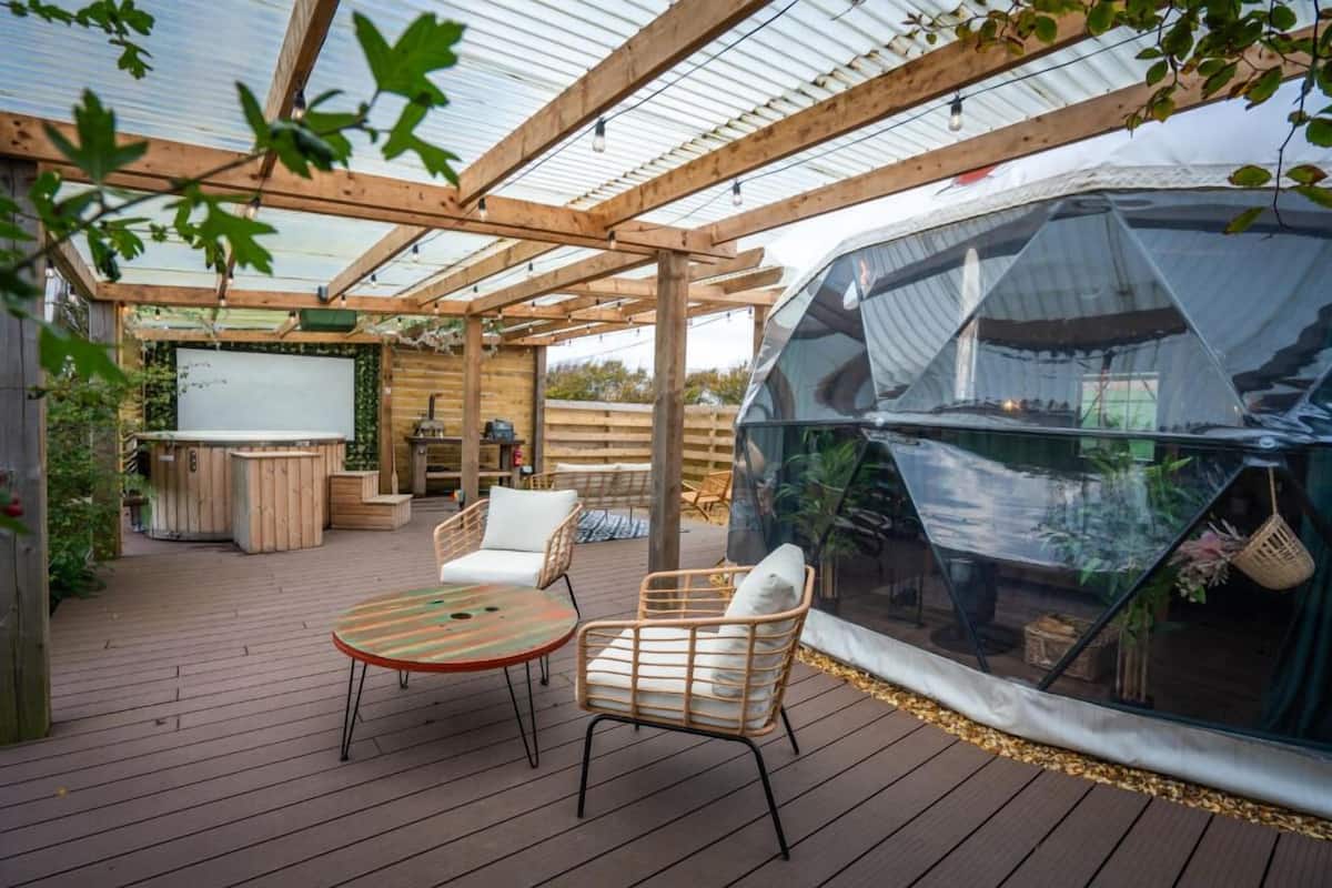 Un espacio al aire libre techado con una terraza de madera, una estructura de invernadero de vidrio y una zona de estar con una mesa y sillas.