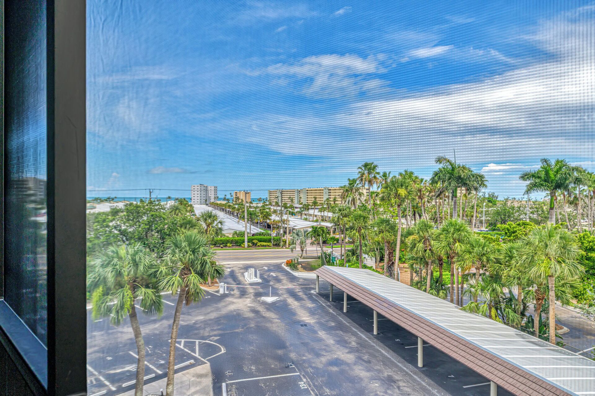 A parking area with a covered walkway, palm trees, and buildings in the background.