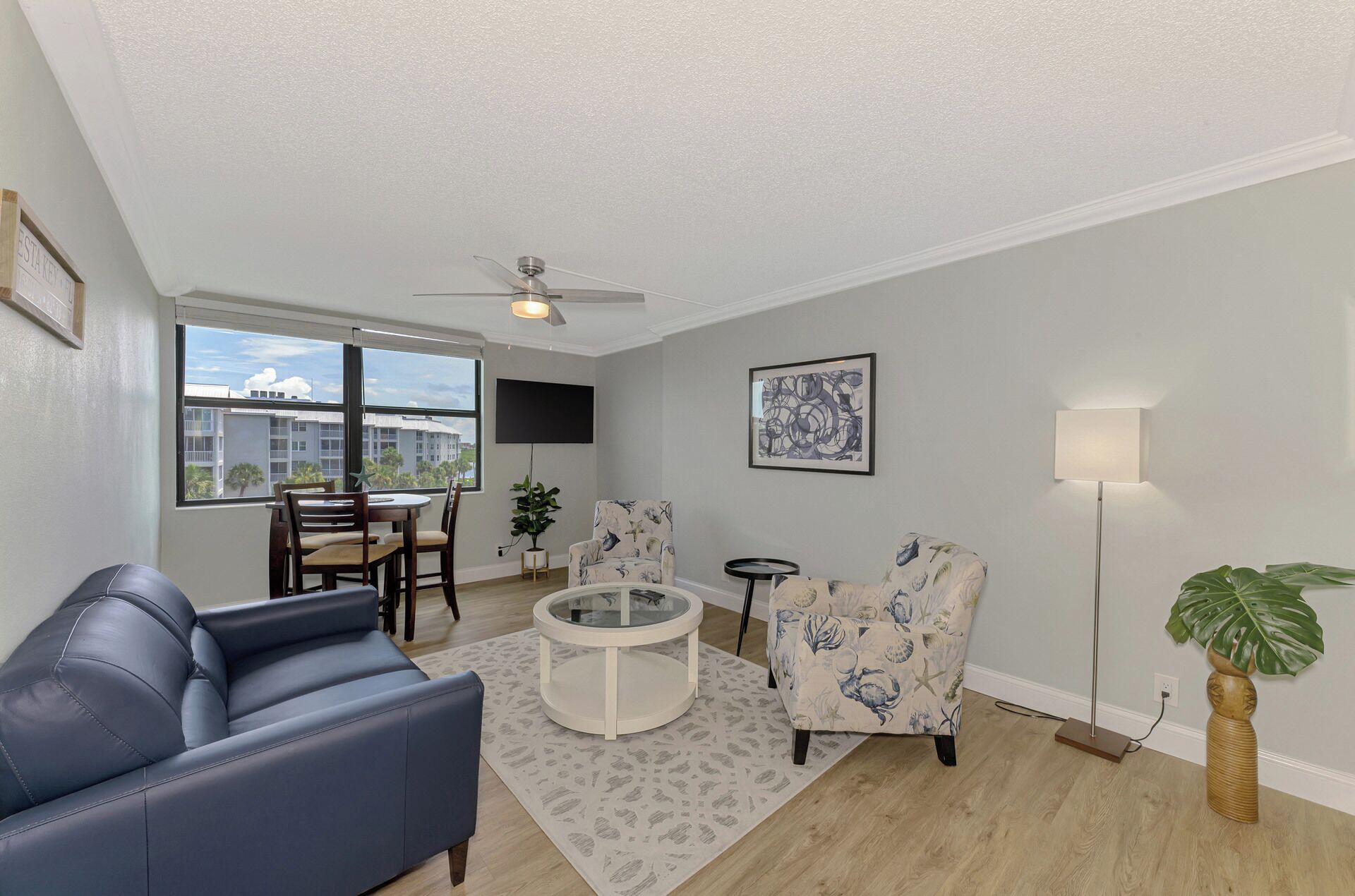 A modern living room with a blue sofa, a round coffee table, and two patterned armchairs. A ceiling fan and a TV are mounted on the wall.