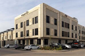 Un edificio de varios pisos con fachada plana, múltiples ventanas y un estacionamiento con varios vehículos.