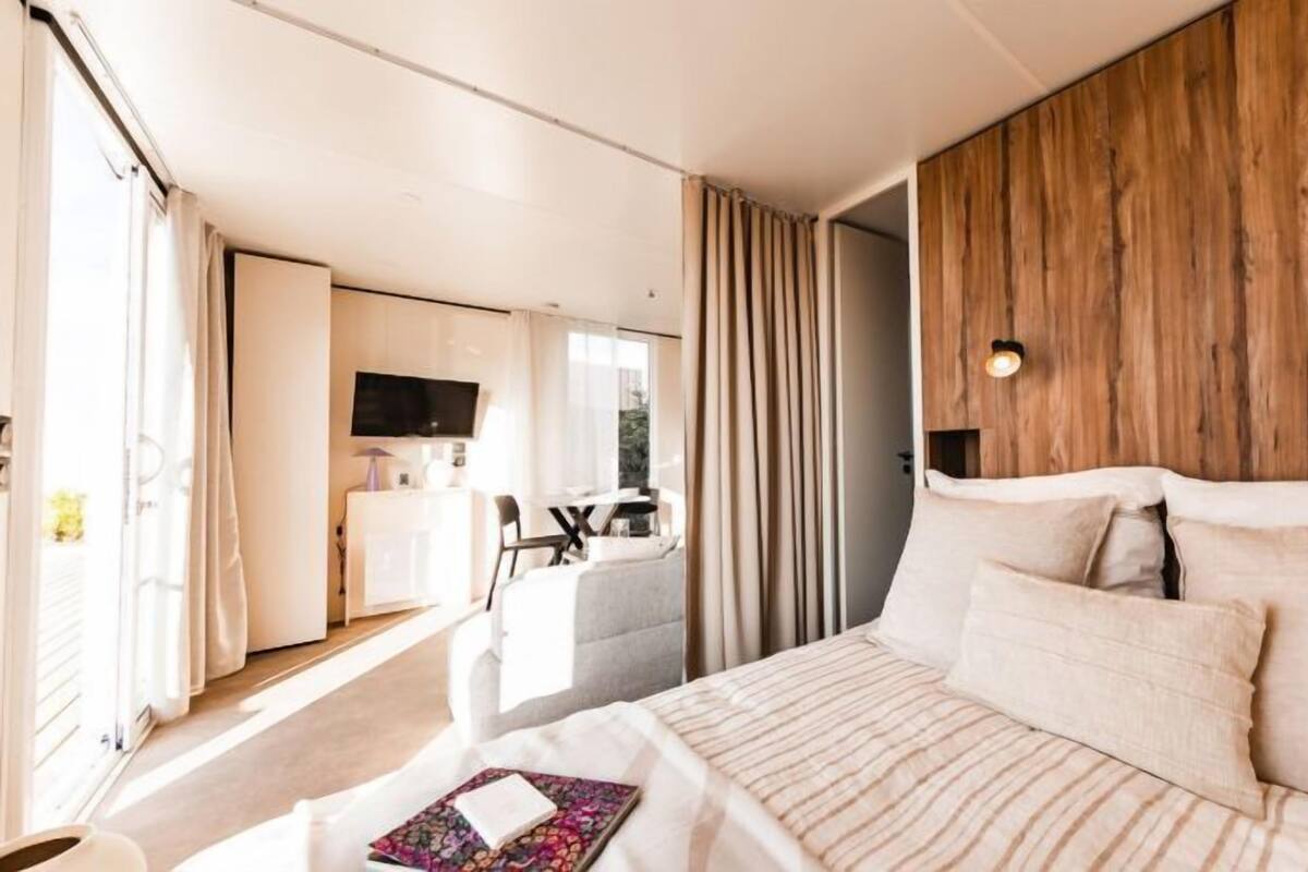 Ein modernes Hotelzimmer mit einem großen Bett, einer Wand mit Holzvertäfelung, einem Fernseher und einem Essbereich, der durch eine offene Tür sichtbar ist.