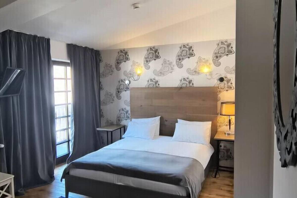 Ein Hotelzimmer mit Bett, Nachttisch, Lampe, Fenster mit Vorhängen und einer Wand mit einem Blumenmuster.