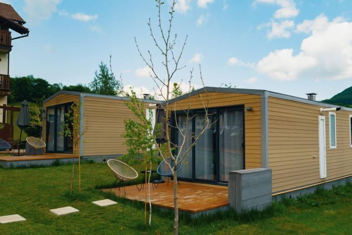 Ein modernes Mobilheim mit Terrasse, einem kleinen Baum und einer Satellitenschüssel.