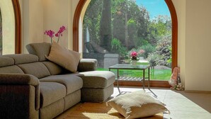 A modern living room with a sectional sofa, a small table, and a view of a garden through a large arched window. - Villa in the Sibillini Mountains, Pool & Privacy (Sant'Angelo in Pontano)