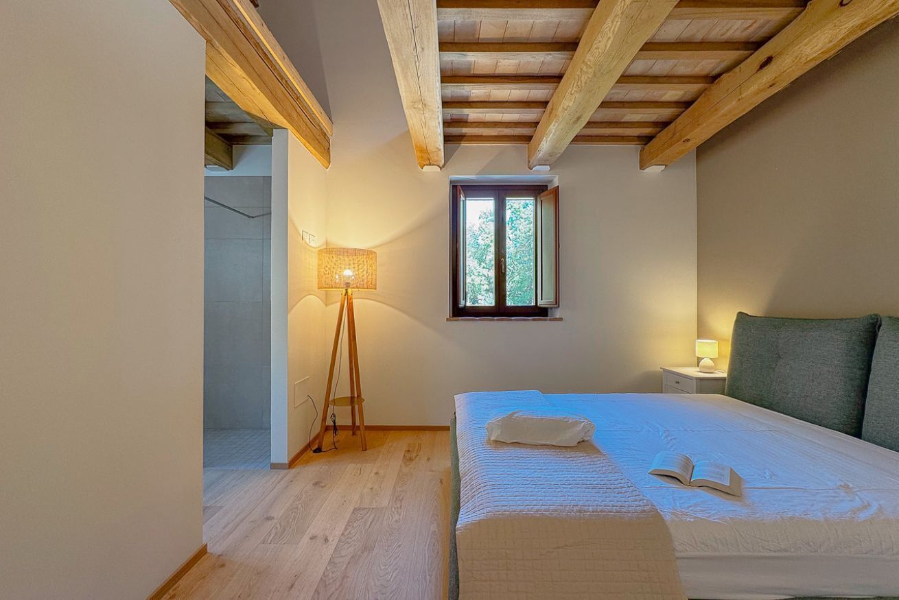 A bedroom with a wooden ceiling, a bed with a white duvet, a bedside lamp, and a window with a view of trees.
