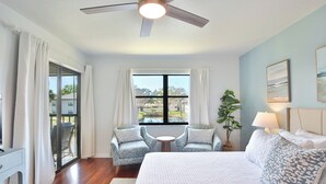 Ein Schlafzimmer mit Bett, Deckenventilator, Fenster mit Vorhängen, kleinem Tisch, zwei Stühlen und einer Pflanze.