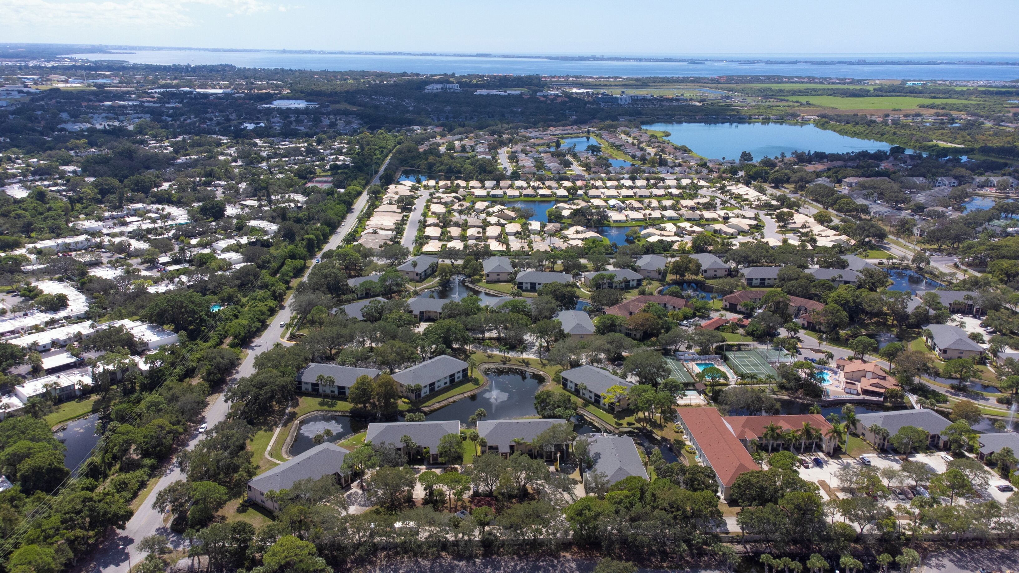 An aerial view of a residential area with multiple houses, a central swimming pool, and a large body of water in the background.