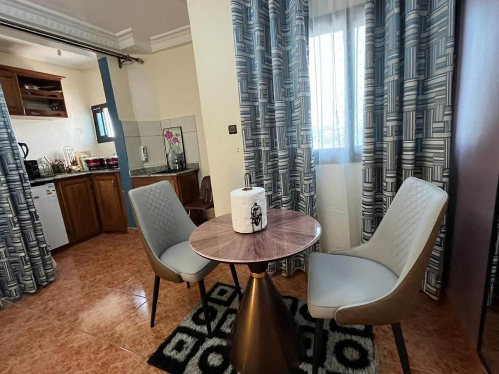 Un comedor con dos sillas y una mesa redonda, una cocina al fondo y cortinas con estampado.