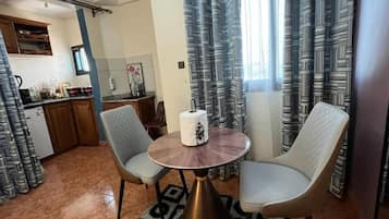 Un comedor con dos sillas y una mesa redonda, una cocina al fondo y cortinas con estampado.