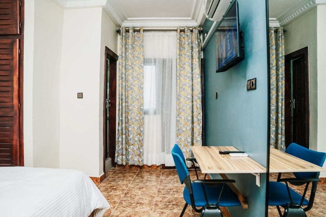 A room with a bed, a dining table with chairs, a TV mounted on the wall, and a window with curtains.