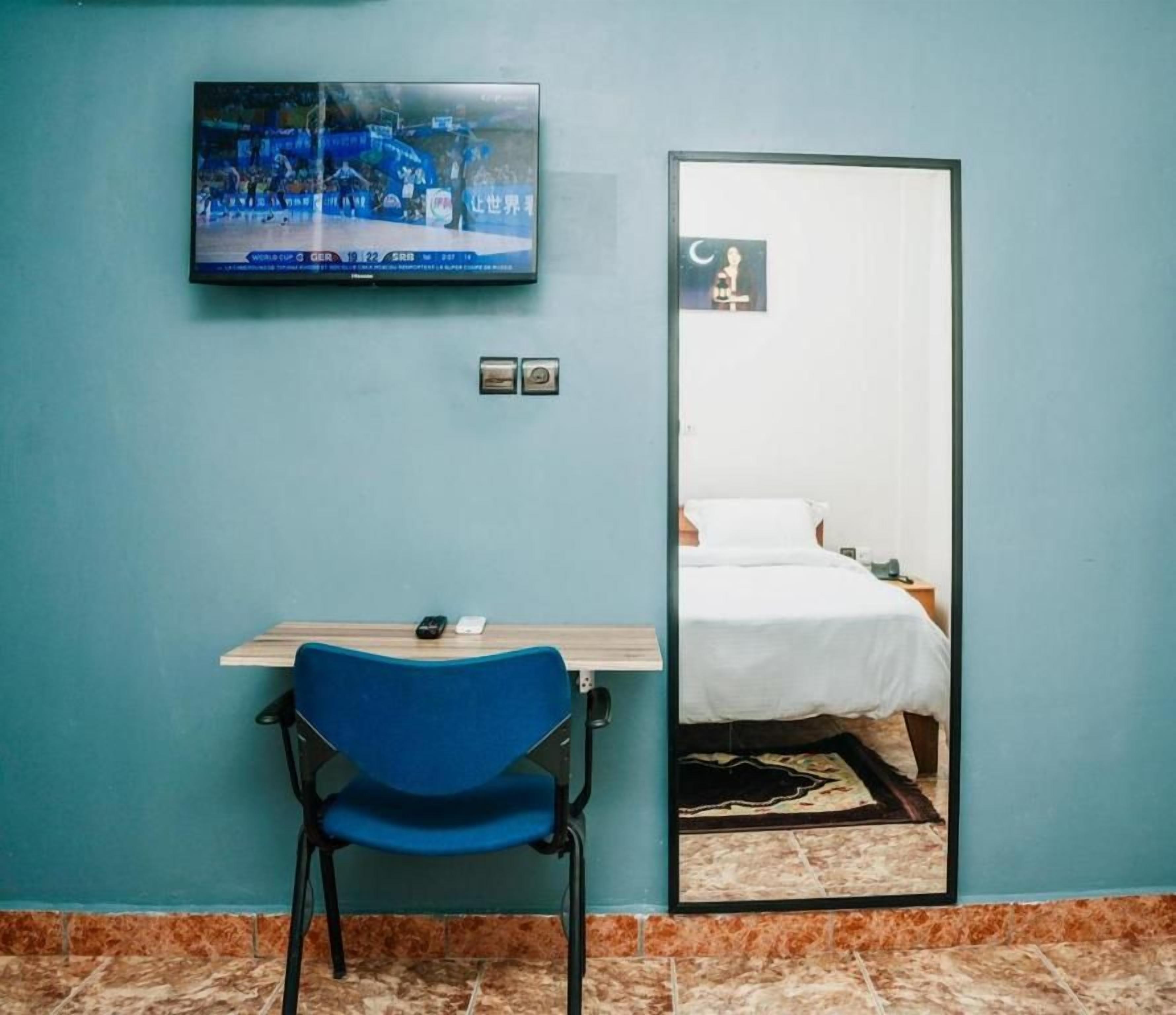 Una habitación con cama, escritorio con silla, televisor montado en la pared y un espejo que refleja la cama.