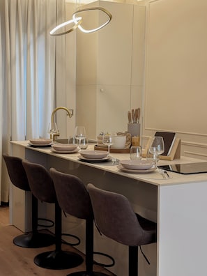 Un espace repas moderne avec une longue table, trois tabourets de bar, un luminaire suspendu et une tablette posée sur la table.
