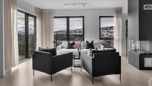 A modern living room with a sofa, two armchairs, a coffee table, and a view of a cityscape through large windows. - TotalApartments Gjøa Premium (Tromsø)