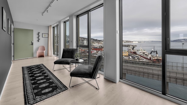 A spacious room with large windows, a black rug, two chairs, and a view of a harbor. - TotalApartments Gjøa Premium (Tromsø)