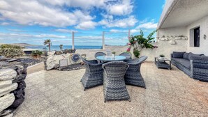 An outdoor patio with wicker furniture, a table, and a sofa, overlooking a coastal landscape. - Casa Buenavista • Pool • Ocean views • Beach 2 min • LVH (Famara)