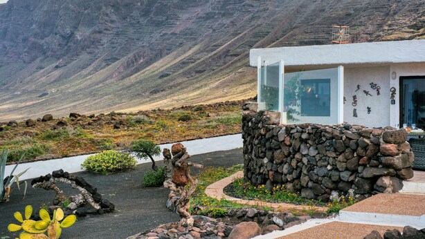 A modern building with a stone wall, a glass door, and a view of a mountainous landscape. - Casa Buenavista • Pool • Ocean views • Beach 2 min • LVH (Famara)