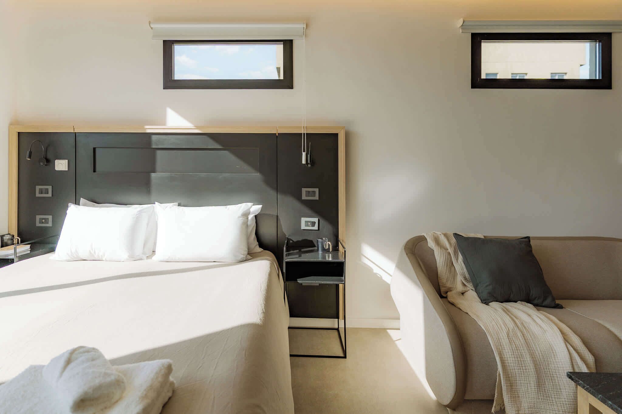 A hotel room with a bed, a sofa, a small table, and two windows.