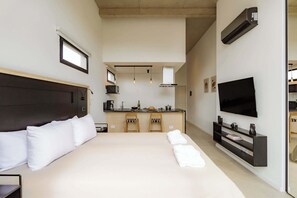 Ein modernes Hotelzimmer mit einem großen Bett, einem Fernseher, einer Küchenzeile und Blick zur Küche.