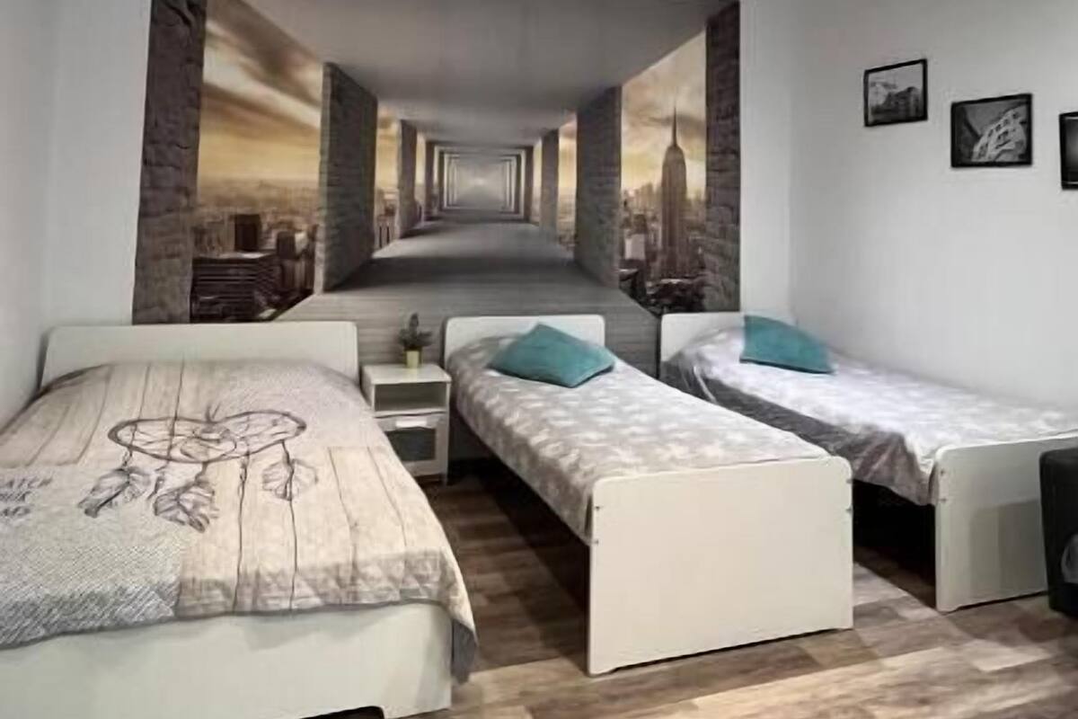 A modern bedroom with a large bed, two smaller beds, a mirror, and a cityscape mural on the wall.