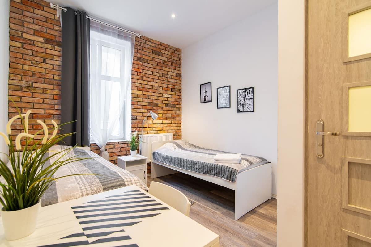 A bedroom with a brick wall, a bed, a bedside table, a lamp, and a window with curtains.