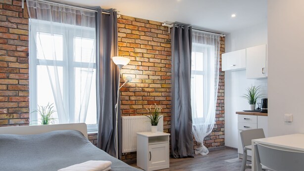 Una habitación moderna con pared de ladrillo, una cama con manta gris, una pequeña mesa blanca con una planta, un armario blanco y una zona de kitchenette.