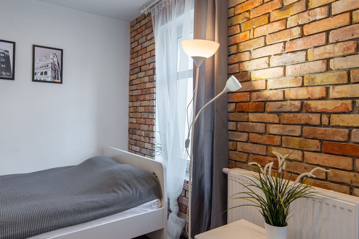 A bedroom with a brick wall, a bed, a floor lamp, and a potted plant.