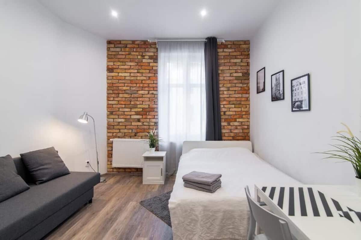 A modern bedroom with a brick accent wall, a bed, a sofa, a small table, and a radiator.