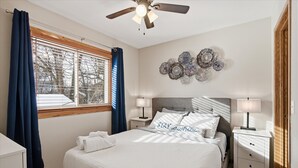 Une chambre à coucher comprenant un lit, deux tables de chevet, un ventilateur de plafond, une fenêtre avec des rideaux et des éléments de décoration murale.