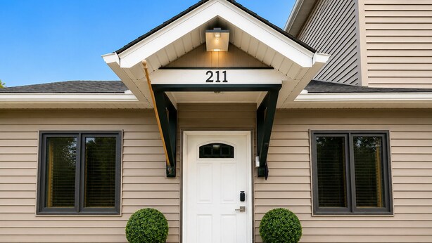 Une maison avec une porte blanche, le numéro 211, deux plantes en pot et un paillasson noir.
