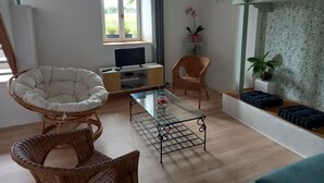 Een woonkamer met een glazen salontafel, rieten stoelen, een televisie op een houten kast en uitzicht op groen buiten.