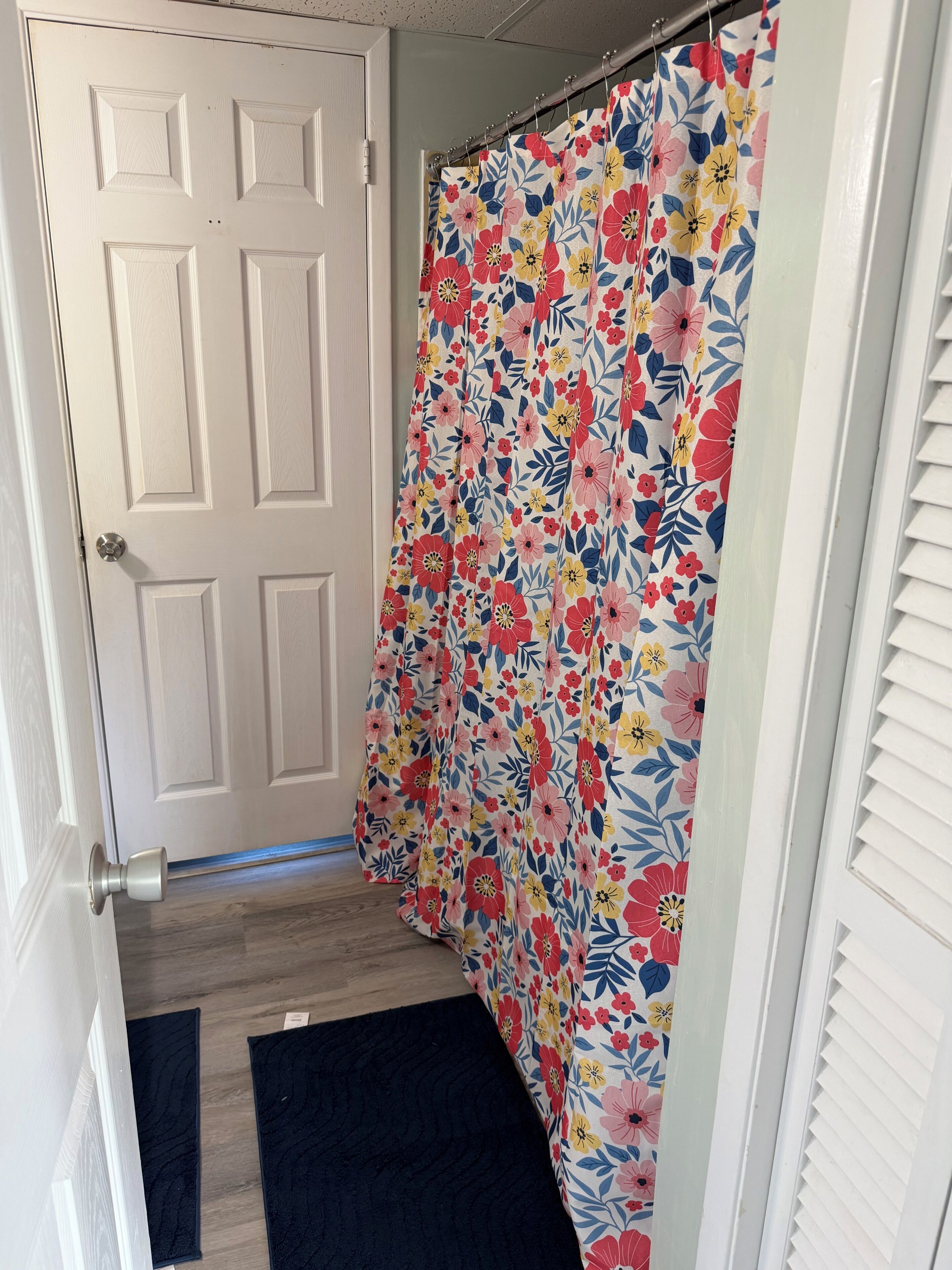 A bathroom with a white door, a shower curtain with a floral pattern, a dark blue rug, and a white window with blinds.