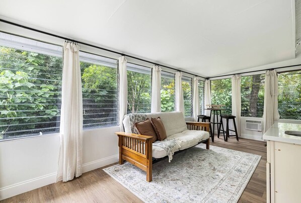 Une pièce confortable avec un canapé, une petite table et une chaise. De grandes fenêtres, ornées de rideaux, offrent une vue sur la verdure environnante.