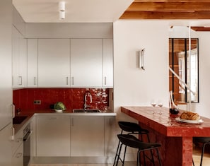 A modern kitchen with white cabinets, a red backsplash, and a wooden ceiling. There is a bar area with stools and a bottle of wine. - Apartment of 78 m² in Neuilly-sur-Seine (Neuilly-sur-Seine)