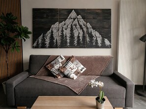 Una sala de estar moderna con un sofá gris, una mesa de centro de madera y una obra de arte de gran tamaño con temática de montaña en la pared.