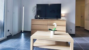 Ein modernes Wohnzimmer mit einem Flachbildfernseher auf einer Holzkommode, einem kleinen Tisch mit einer Topfpflanze und einem großen Fenster.
