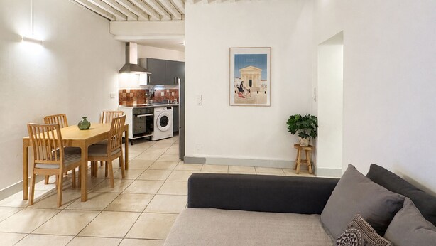 Ein Wohnzimmer mit einer Couch, einem Esstisch mit Stühlen, einer Waschmaschine und einer Küche im Hintergrund.