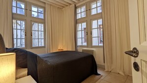 Ein Schlafzimmer mit Bett, Nachttisch, Lampe und Fenstern mit Vorhängen.