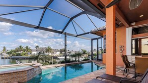 Zona de piscina con cerramiento de vidrio, sillones de descanso y vista a un lago y casas.