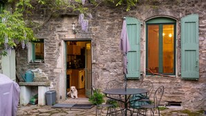 Ein stattliches Steinhaus mit rustikalem Charme, mit einer kleinen Außenbestuhlung mit Metallstühlen und einem Tisch, einem draußen sitzenden Hund und einer im Inneren sichtbaren Waschmaschine.