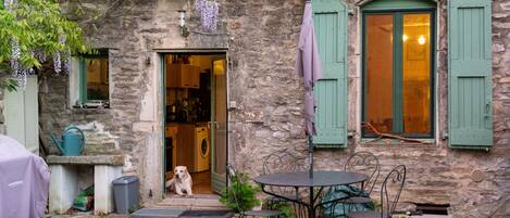 Un edificio de piedra con encanto rústico, que cuenta con una pequeña zona de estar al aire libre con sillas y una mesa de metal, un perro sentado afuera y una lavadora visible en el interior.