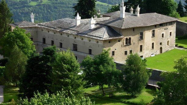 A historic building with multiple chimneys, surrounded by lush greenery and a mountainous backdrop.