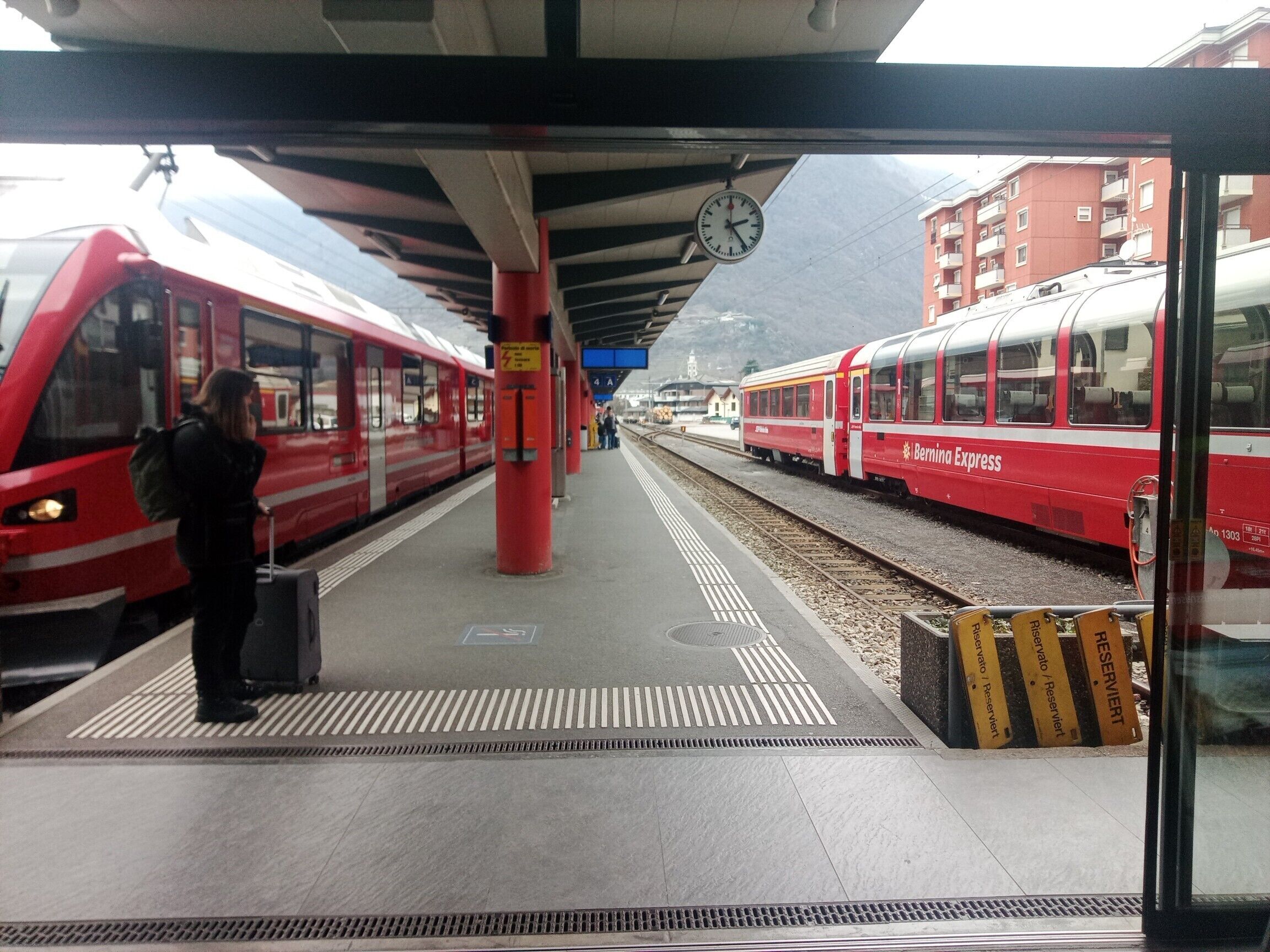 Un quai de gare avec deux trains rouges, une horloge et une personne portant des bagages.